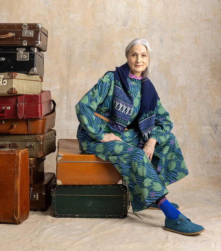 A woman sitting on a stack of suitcases wearing layers of clothing from Gudrun Sjödén.