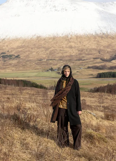 A woman standing in a mountain landscape wearing a wool coat and knitted accessories from Gudrun Sjödén.