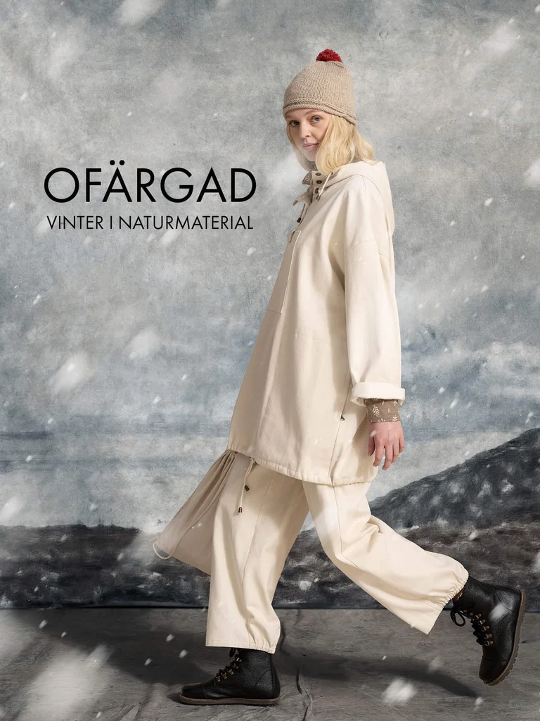A woman walking while snow falls, wearing a knit hat, a white anorak and a pair of white pants from Gudrun Sjödén.