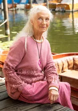 Eine Dame sitzt am Bootssteg mit roséfarbenen Pullover. Der untere Teil des Oberteils hat Blumen aufgestickt. Untenrum trägt die Dame einen gestreiften Rock in altrosé.
