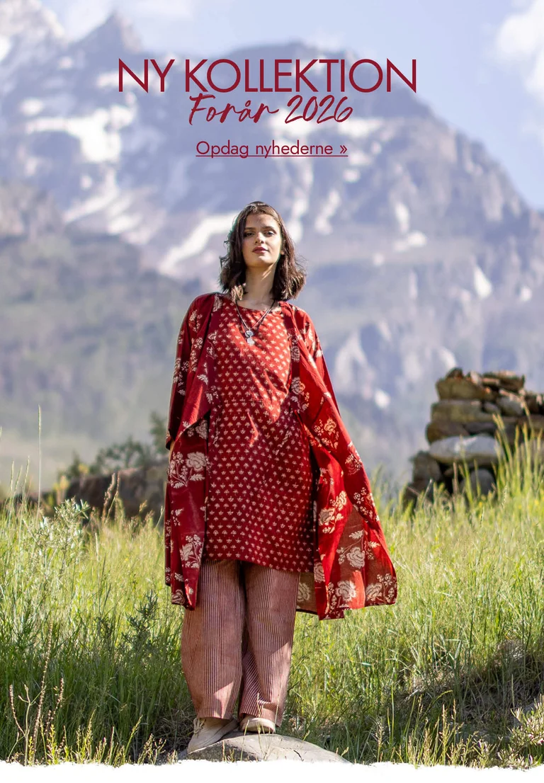 En kvinde, der står i et bjergrigt landskab, klædt i lag på lag af rødt og vævet tøj fra Gudrun Sjödén.