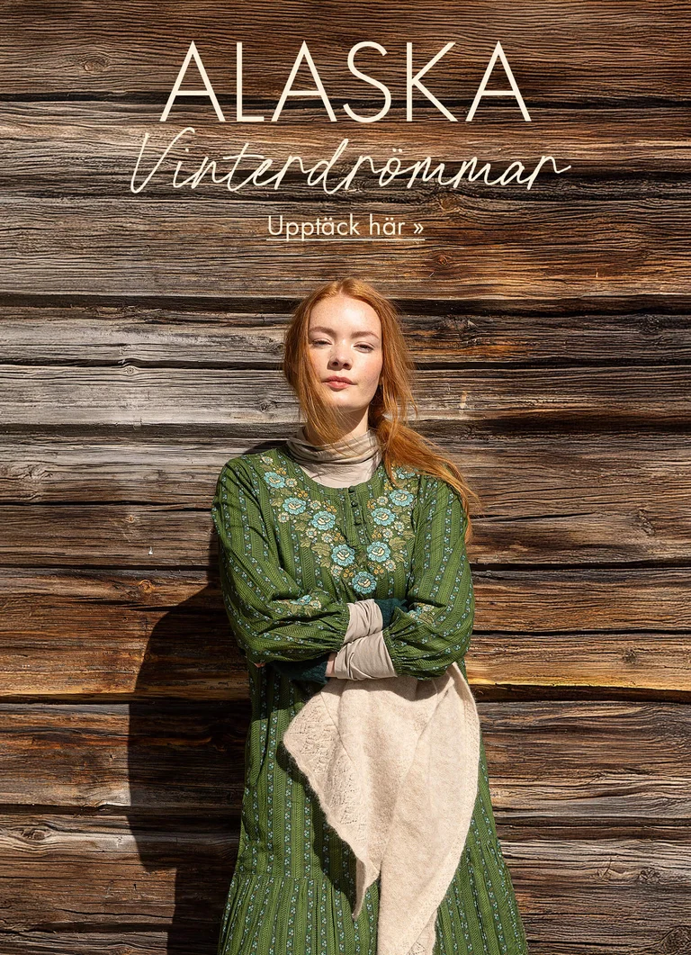 En kvinna som står mot en trävägg med armarna i kors iklädd en grön klänning med broderier från Gudrun Sjödéns julkollektion 2025. 