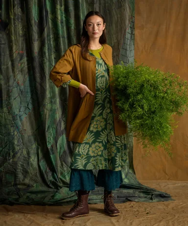 A woman holding a bunch of green leaves wearing layers of clothes in earthy hues from Gudrun Sjödén.