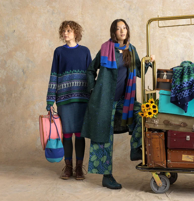 Two women standing beside a luggage cart, wearing layers of clothing from Gudrun Sjödén.