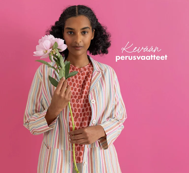 A woman standing against a pink background wearing essentials from Gudrun Sjödén’s 2026 Spring collection. Holding a pink flower in her hand.