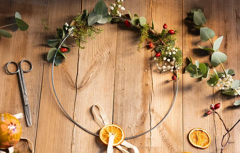 A Christmas wreath on a wooden table with scissors, greenery and dried oranges.