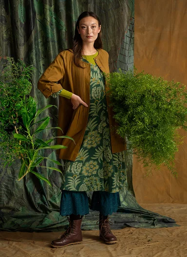 En kvinde som holder en stor grøn plante og er iført lag på lag af tøj fra Gudrun Sjödén.