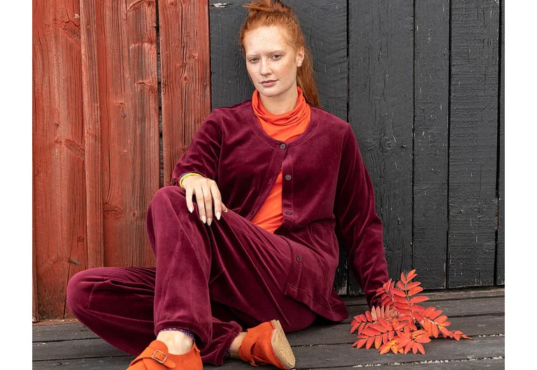 A woman sitting on a step, next to a cluster of rowan berries, wearing a burgundy velour cardigan and burgundy velour trousers over an orange polo-neck top from Gudrun Sjödén.