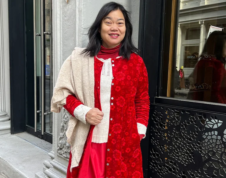 Suelain Moy wearing layered red and cream Gudrun Sjödén clothing, smiling outside an ornate city building.