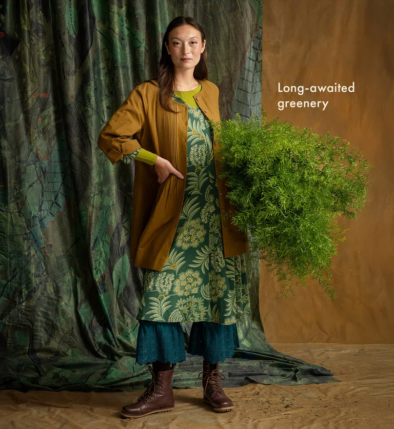 A woman holding a green plant, wearing layers of garments from Gudrun Sjödén.