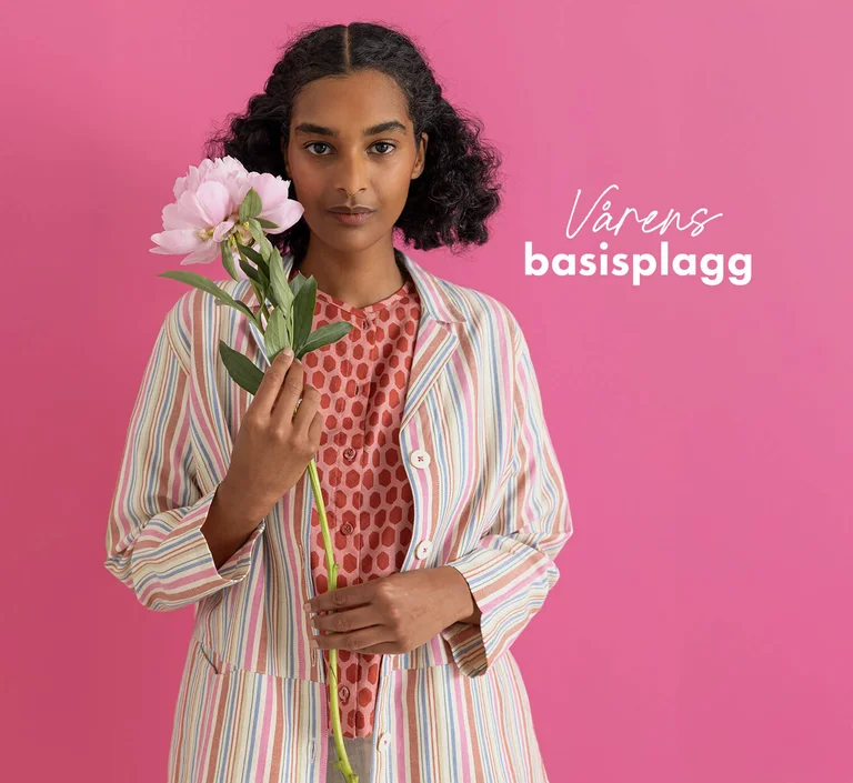 A woman standing against a pink background wearing essentials from Gudrun Sjödén’s 2026 Spring collection. Holding a pink flower in her hand.