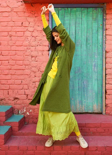 A woman standing in front of a turquoise door wearing the knitted long cardigan ”Misty” from Gudrun Sjödén.