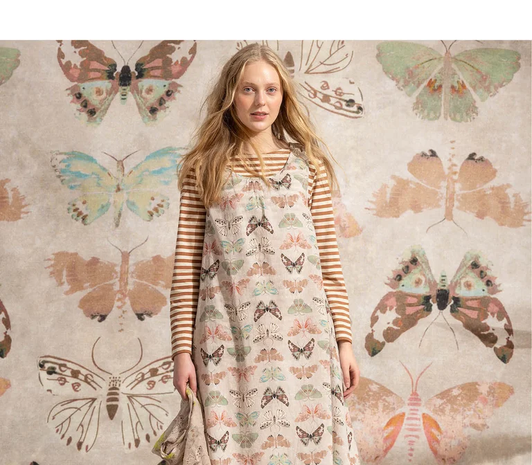 A woman standing against a butterfly background wearing a beige dress with a butterfly pattern from Gudrun Sjödén.