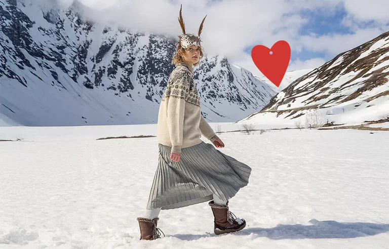 A woman with an animal mask, walking in a snowy landscape, wearing a beige and grey patterned, knitted sweater from Gudrun Sjödén’s 2025 Christmas collection. There is also a red, graphic heart in the picture.