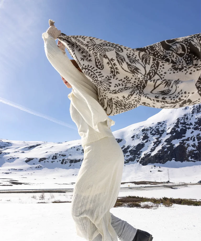 A woman running in a snowy landscape with a blanket from Gudrun Sjödén’s 2025 Christmas collection over her head.