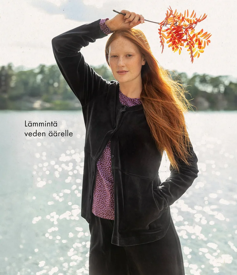 A woman in front of water wearing a black velour cardigan with matching velour pants from Gudrun Sjödén. She is holding a branch from a mountain ash tree in her hand.