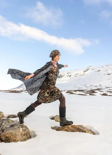 A woman with an animal mask on her head wearing a red and patterned outfit with a knitted cardigan from Gudrun Sjödén’s Alaska’s Wild Heart collection.