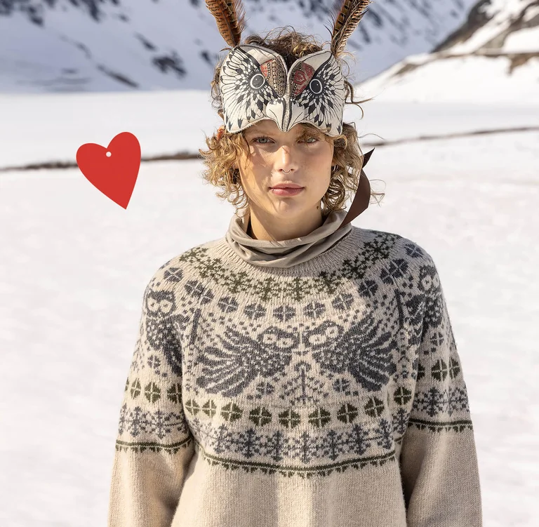 A woman with an animal mask wearing a beige and grey patterned, knitted sweater from Gudrun Sjödén’s 2025 Christmas collection. There is also a red, graphic heart in the picture.