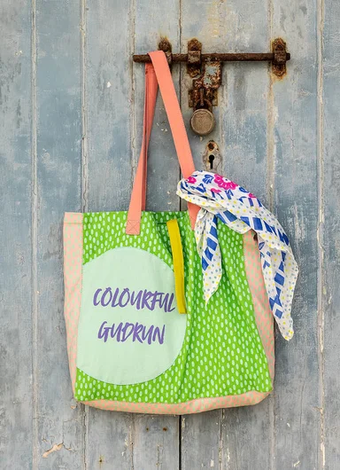 A green and orange patterned bag decorated with the words “Colourful Gudrun” and hanging on a wooden door.