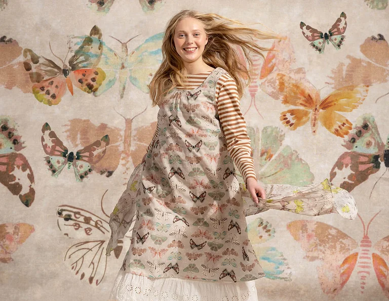 A woman standing against a butterfly background wearing a beige dress with a butterfly pattern from Gudrun Sjödén.