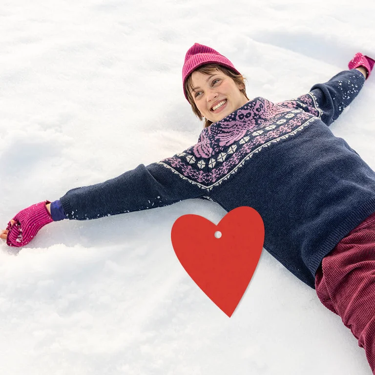 A woman lying on the snow laughing, wearing Luna sweater with its owl pattern from Gudrun Sjödén’s Christmas collection. There is also a red, graphic heart in the picture.