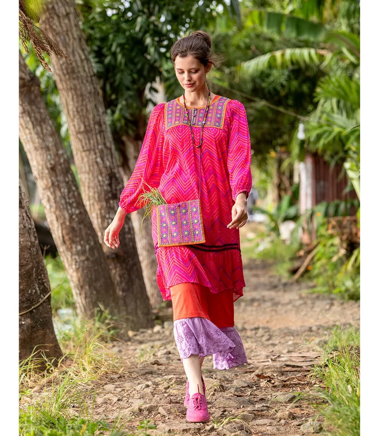 A woman walking along a path surrounded by green trees, wearing layers of pink and purple clothes from Gudrun Sjödén.