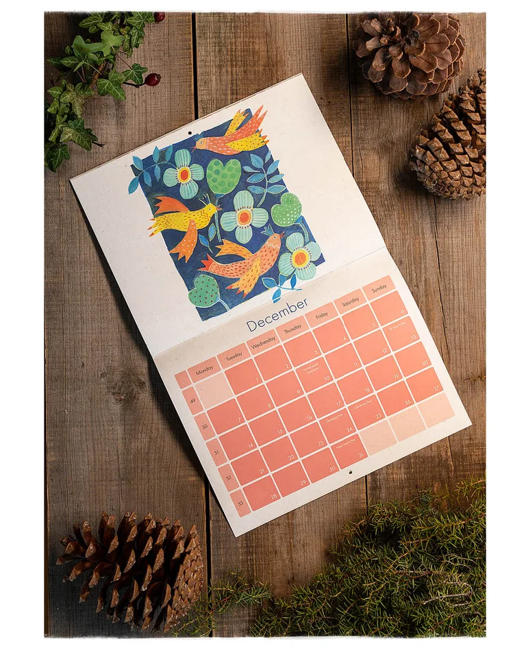 A calendar with watercolours from Gudrun Sjödén lying next to cones and greenery on a wooden surface.