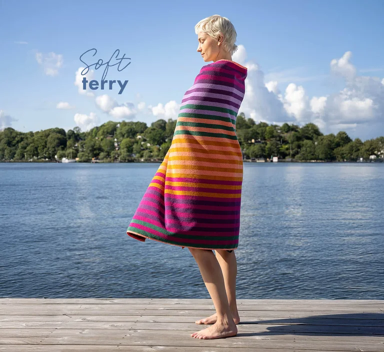 A woman standing on a jetty looking out over the water wrapped in a striped, multicoloured terry towel from Gudrun Sjödén.