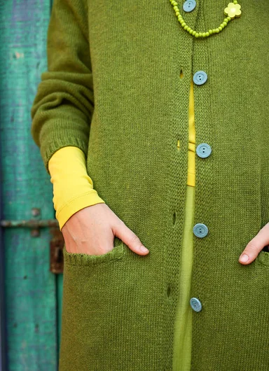 A woman standing in front of a turquoise door wearing the knitted long cardigan ”Misty” from Gudrun Sjödén.