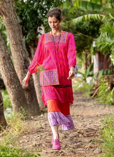 A woman wearing a pink, patterned dress and a small, pink bag adorned with mirrors from Gudrun Sjödén.