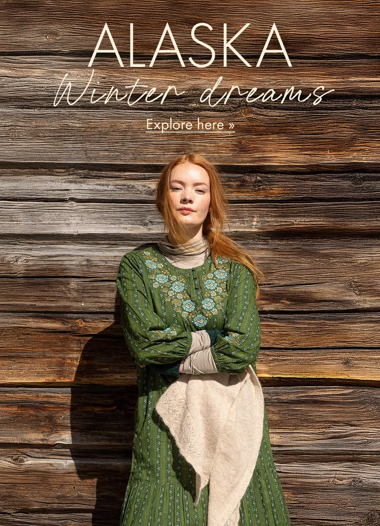A woman standing against a timber wall with her arms crossed wearing a green dress with embroidery from Gudrun Sjödén’s 2025 Christmas collection.