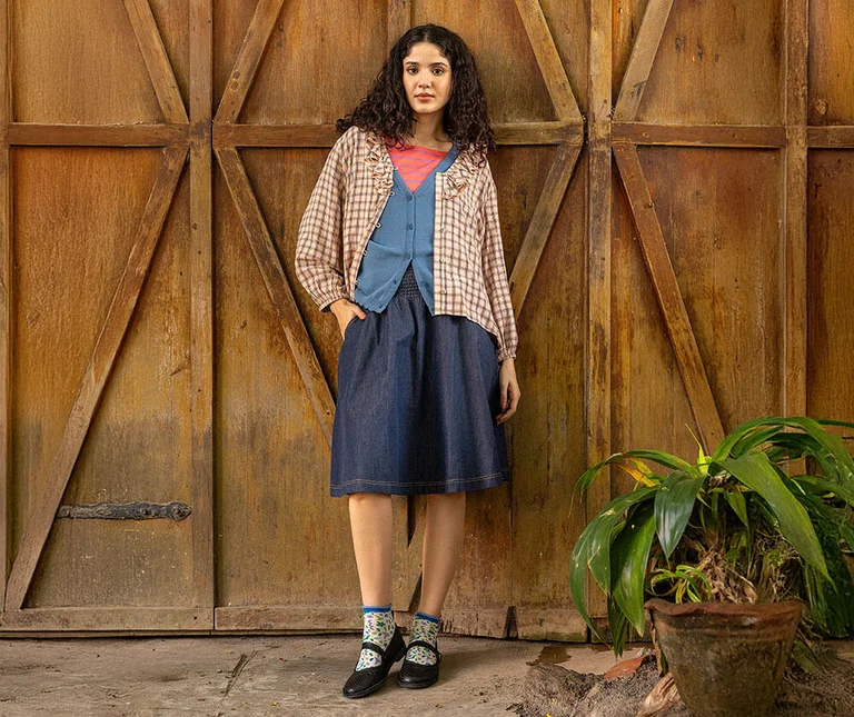 A woman leaning against a wall wearing a ruffle blouse matched with a blue cardigan and jeans skirt from Gudrun Sjödén.